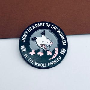May include: A circular enamel pin with a black background, displaying a cartoon opossum. The opossum is grey, white, and pink, with its tongue out. The text reads "DON'T BE A PART OF THE PROBLEM BE THE WHOLE PROBLEM." Two small bin icons are present.