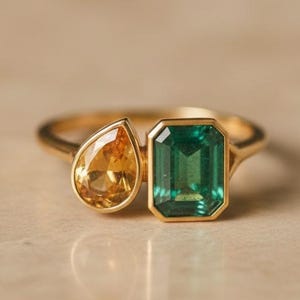 May include: A gold ring featuring two gemstones: a pear-shaped yellow gemstone and an emerald-cut green gemstone. The ring has a delicate band and a modern design. The gemstones are set side by side, creating a unique and eye-catching look.