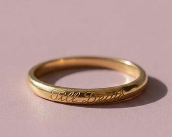 May include: A polished gold band ring featuring the engraved script text Till Death on the outer surface. The ring has a smooth, rounded profile and a reflective metallic finish, showcasing a simple and classic design with clear lettering.