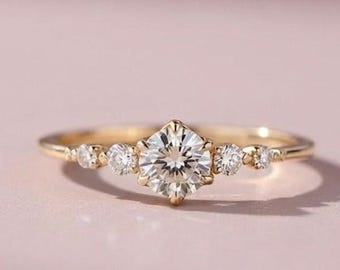 May include: A gold ring featuring a large round brilliant-cut center stone flanked by two smaller round stones on each side, set in a delicate polished gold band. The design showcases a total of five clear stones arranged in a horizontal row.