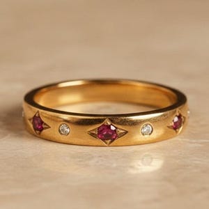 May include: A gold ring featuring alternating ruby and diamond embellishments. The rubies are set in a star-shaped pattern, with round diamonds interspersed. The ring has a polished finish and is presented on a neutral background.