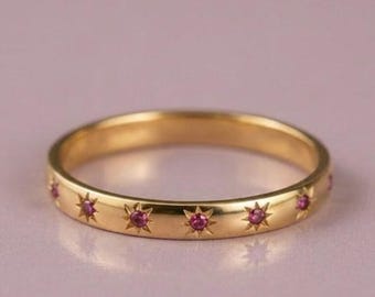 May include: A gold band ring featuring a series of small, evenly spaced star-shaped engravings, each centered with a tiny, vibrant pink gemstone. The metal has a polished, reflective finish, and the design wraps continuously around the ring.
