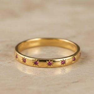 May include: A gold ring with a series of starburst designs, each containing a small, round, red gemstone. The ring is set against a neutral background, highlighting its polished, reflective surface. The ring is a piece of jewelry.