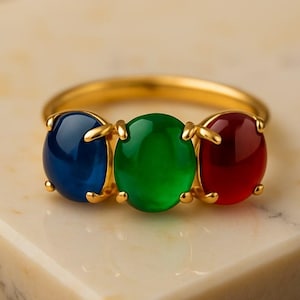 Könnte beinhalten: Ein goldener Ring mit drei ovalen Edelsteinen in Blau, Grün und Rot. Die Edelsteine sind in einem goldenen Band gefasst und bilden ein auffälliges Schmuckstück. Der Ring wird auf einer Marmoroberfläche präsentiert.