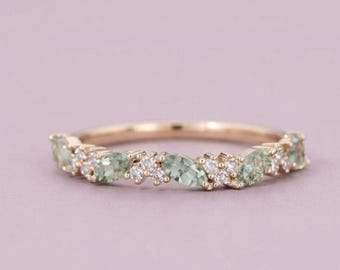 May include: A rose gold ring featuring a series of marquise-cut light green stones interspersed with small, round, clear white stones arranged in a cluster along the top half of the thin, polished metal band.
