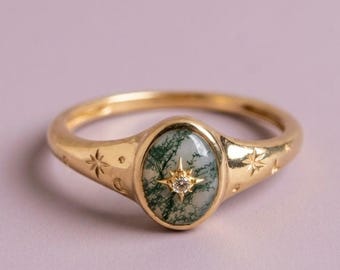 May include: A gold ring featuring an oval moss agate stone set in the center. The stone is accented with a small, star-shaped setting containing a tiny clear gemstone. The gold band is engraved with small star and crescent moon motifs.
