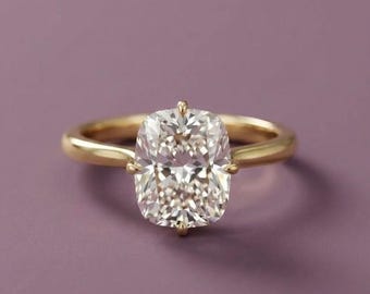 May include: A gold ring featuring a large, clear, cushion-cut gemstone set in a four-prong setting. The band is smooth and polished, showcasing a classic design with a central stone that reflects light with multiple facets across its surface.