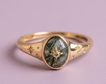 May include: A gold ring featuring an oval moss agate stone with a central star-shaped setting containing a small clear gemstone. The gold band is engraved with small star and dot motifs on both sides of the central oval stone setting.