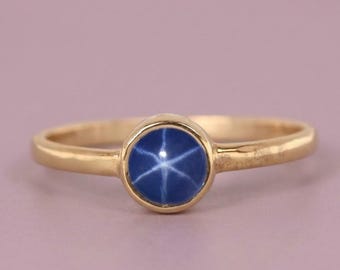 May include: A gold ring featuring a round, cabochon-cut blue star sapphire gemstone set in a simple bezel. The gemstone displays a distinct six-rayed star pattern across its surface, and the band is crafted from a smooth, polished yellow gold metal.