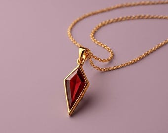 May include: A gold necklace featuring a kite-shaped pendant with a deep red gemstone set in a polished gold frame. The pendant hangs from a delicate gold chain, showcasing a sharp, geometric design with clean lines and a vibrant, translucent red center.