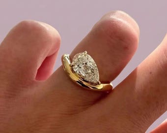 May include: A gold ring featuring a single pear-shaped diamond set in a smooth, polished yellow gold band. The diamond is positioned at an angle, secured by a prong setting that rests against the curved, thick band of the ring.