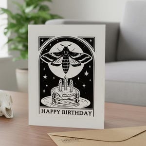 May include: A white birthday card featuring a black and white design of a moth above a cake with candles. The words "HAPPY BIRTHDAY" are printed below. A brown envelope sits on a wooden table.