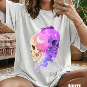May include: A light gray t-shirt featuring a graphic of two colorful skulls. One skull is yellow and pink, the other is purple. Both have crescent moon details. The word "WHITE" is printed in a stylized font at the bottom.