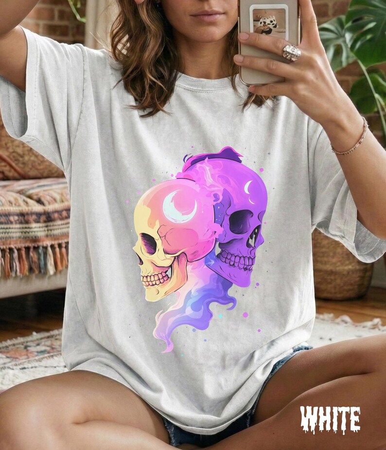 May include: A light gray t-shirt featuring a graphic of two colorful skulls. One skull is yellow and pink, the other is purple. Both have crescent moon details. The word "WHITE" is printed in a stylized font at the bottom.