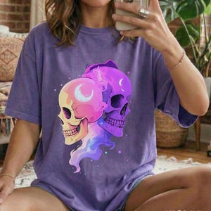 May include: A violet-colored t-shirt featuring a graphic of two stylized skulls. One skull is yellow and pink, the other is purple. Both have crescent moon details. The word "VIOLET" is printed in white at the bottom.