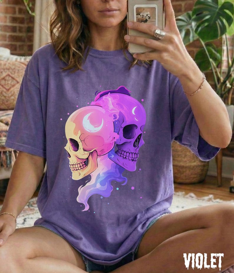 May include: A violet-colored t-shirt featuring a graphic of two stylized skulls. One skull is yellow and pink, the other is purple. Both have crescent moon details. The word "VIOLET" is printed in white at the bottom.