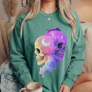 May include: Light green sweatshirt featuring a graphic of two skulls with crescent moon details. One skull is yellow and pink, the other is purple. The text "LIGHT GREEN" is at the bottom.