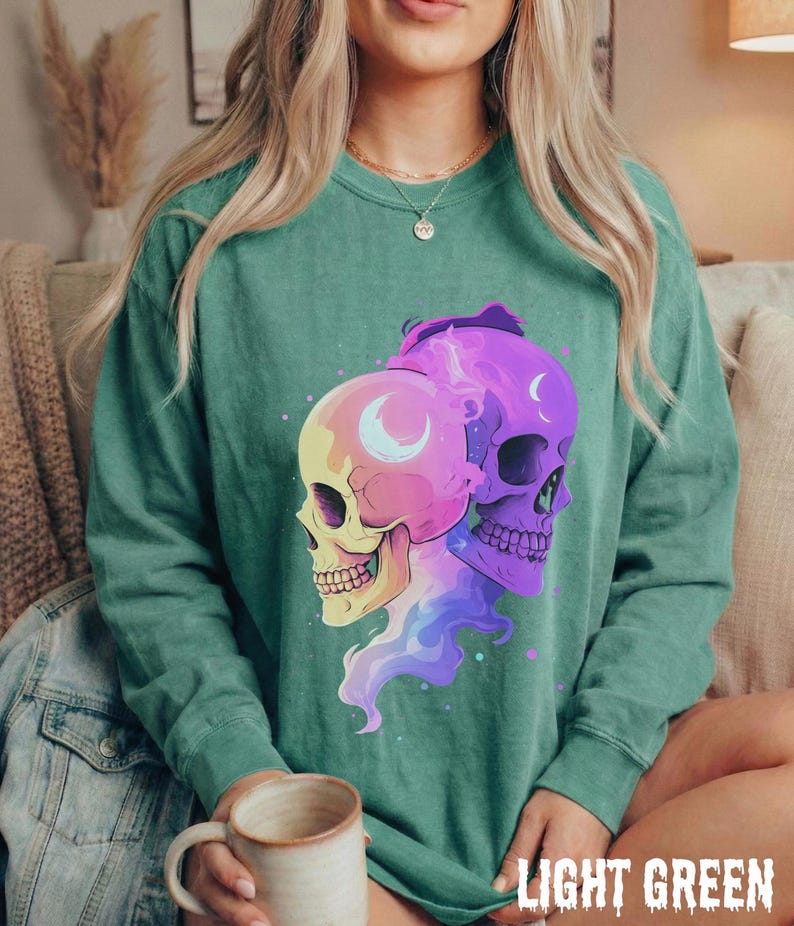 May include: Light green sweatshirt featuring a graphic of two skulls with crescent moon details. One skull is yellow and pink, the other is purple. The text "LIGHT GREEN" is at the bottom.