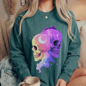 May include: A teal-colored sweatshirt featuring a graphic of two stylized skulls, one yellow and pink, the other purple. Each skull has a crescent moon design. The text "BLUE SPRUCE" is printed in a dripping font at the bottom.