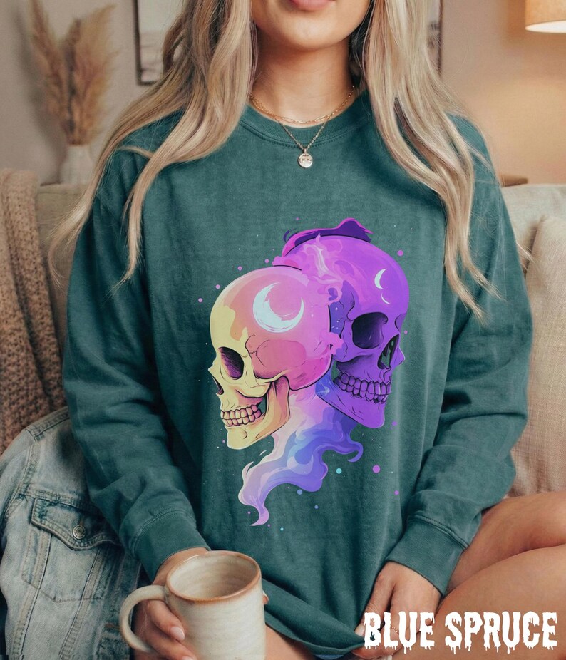 May include: A teal-colored sweatshirt featuring a graphic of two stylized skulls, one yellow and pink, the other purple. Each skull has a crescent moon design. The text "BLUE SPRUCE" is printed in a dripping font at the bottom.