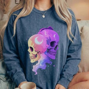 May include: A denim blue long-sleeve sweatshirt featuring a graphic of two stylized skulls. One skull is yellow and pink, the other is purple. Both have crescent moon details. The word "DENIM" is printed in the bottom right corner.