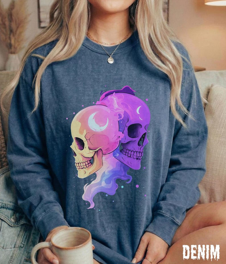 May include: A denim blue long-sleeve sweatshirt featuring a graphic of two stylized skulls. One skull is yellow and pink, the other is purple. Both have crescent moon details. The word "DENIM" is printed in the bottom right corner.