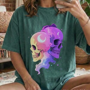 May include: Blue spruce t-shirt featuring a colorful graphic of two skulls with crescent moon details. The skulls are in shades of yellow, pink, and purple, with a flowing, multi-colored design. The text "BLUE SPRUCE" is at the bottom.