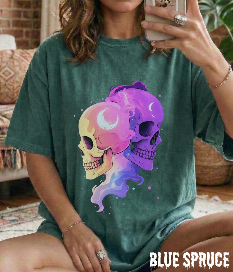 May include: Blue spruce t-shirt featuring a colorful graphic of two skulls with crescent moon details. The skulls are in shades of yellow, pink, and purple, with a flowing, multi-colored design. The text "BLUE SPRUCE" is at the bottom.