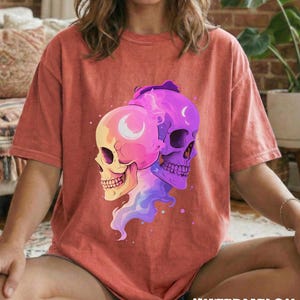 May include: A coral-colored t-shirt featuring a graphic of two stylized skulls, one pink and yellow, the other purple. Each skull has a crescent moon design. The word "WATERMELON" is printed in a distressed font at the bottom.