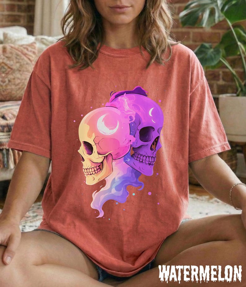 May include: A coral-colored t-shirt featuring a graphic of two stylized skulls, one pink and yellow, the other purple. Each skull has a crescent moon design. The word "WATERMELON" is printed in a distressed font at the bottom.