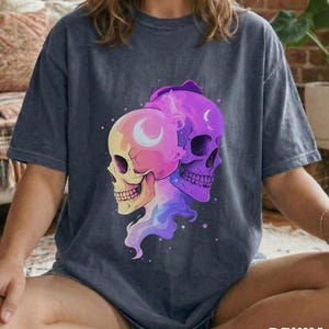 May include: Dark blue t-shirt featuring a colorful graphic of two skulls. One skull is yellow and pink, the other is purple. Both skulls have a crescent moon design. The word "DENIM" is printed at the bottom of the shirt.