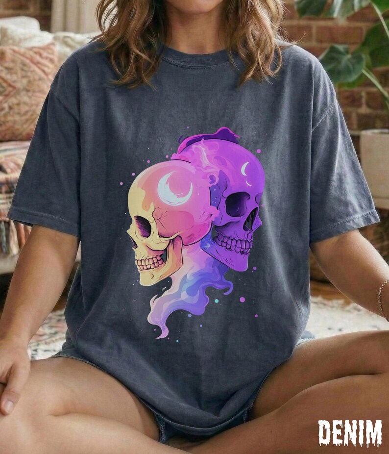 May include: Dark blue t-shirt featuring a colorful graphic of two skulls. One skull is yellow and pink, the other is purple. Both skulls have a crescent moon design. The word "DENIM" is printed at the bottom of the shirt.
