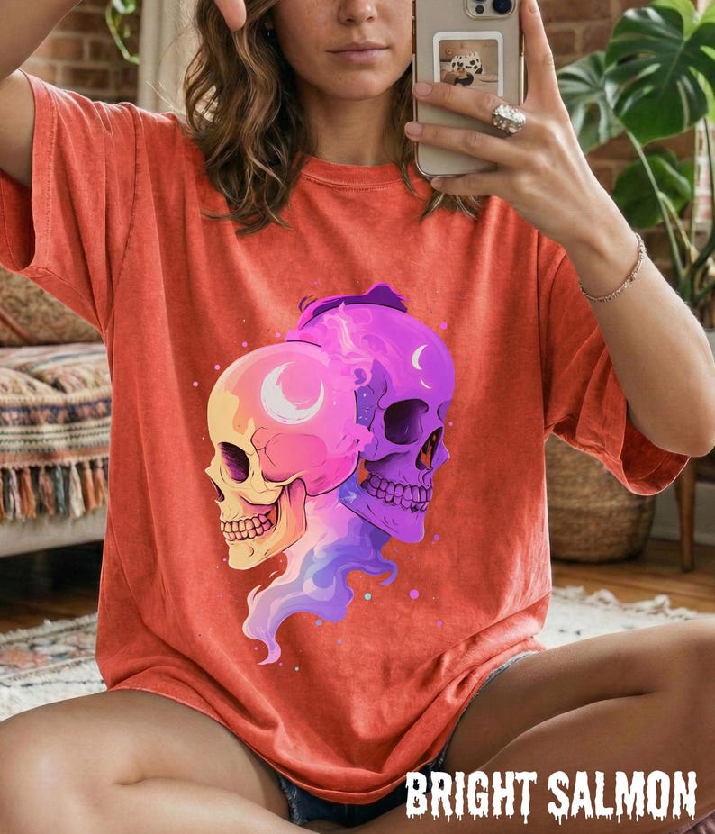 May include: Bright salmon-colored t-shirt featuring a graphic of two stylized skulls. One skull is yellow and pink, the other is purple. The shirt has short sleeves and a relaxed fit. The text "BRIGHT SALMON" is printed at the bottom.
