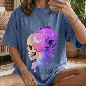 May include: A blue, short-sleeved t-shirt with a graphic of two skulls. One skull is pink and yellow, the other is purple. The graphic includes a crescent moon and a vaporous design. The words "WASHED DENIM" are printed at the bottom.