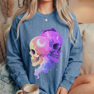 May include: A blue long-sleeve sweatshirt featuring a graphic of two stylized skulls in shades of pink, purple, and yellow. Each skull has a crescent moon design. The text "BLUE JEAN" is printed in a distressed font at the bottom.