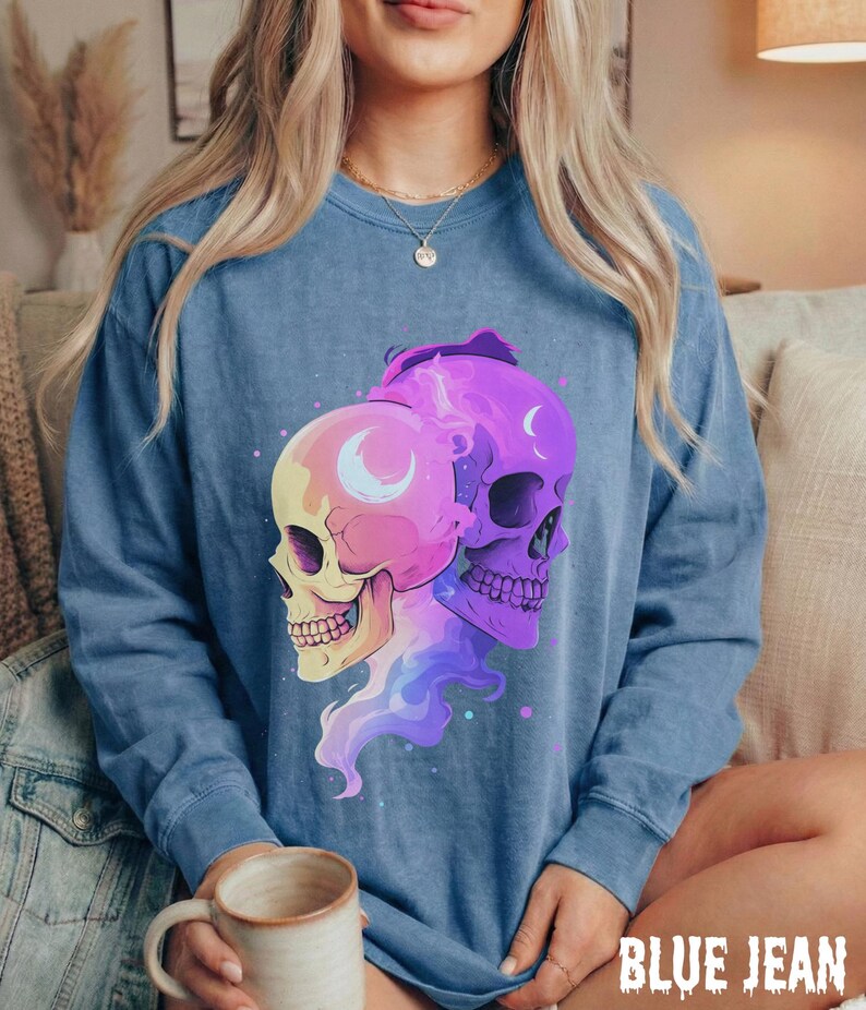May include: A blue long-sleeve sweatshirt featuring a graphic of two stylized skulls in shades of pink, purple, and yellow. Each skull has a crescent moon design. The text "BLUE JEAN" is printed in a distressed font at the bottom.