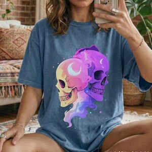 May include: Blue jean colored t-shirt featuring a graphic of two skulls with crescent moon details. The skulls are in shades of yellow, pink, and purple. The text "BLUE JEAN" is at the bottom of the shirt.