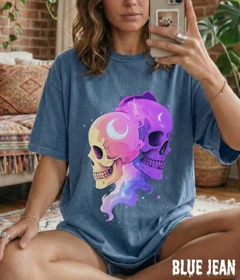 May include: Blue jean colored t-shirt featuring a graphic of two skulls with crescent moon details. The skulls are in shades of yellow, pink, and purple. The text "BLUE JEAN" is at the bottom of the shirt.
