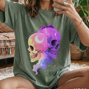 May include: Sage green t-shirt featuring a colorful graphic of two skulls with crescent moon details. The skulls are in shades of pink, purple, and yellow, with a flowing, vaporous design. The word "MOSS" is printed in white at the bottom.