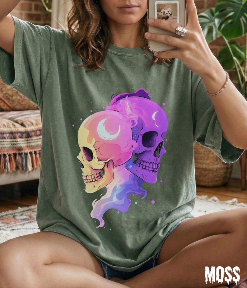 May include: Sage green t-shirt featuring a colorful graphic of two skulls with crescent moon details. The skulls are in shades of pink, purple, and yellow, with a flowing, vaporous design. The word "MOSS" is printed in white at the bottom.