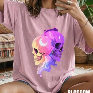 May include: A light pink t-shirt featuring a colorful graphic of two skulls with crescent moon details. The skulls are in shades of yellow, pink, and purple, with a flowing design. The word "BLOSSOM" is printed in a white, stylized font.