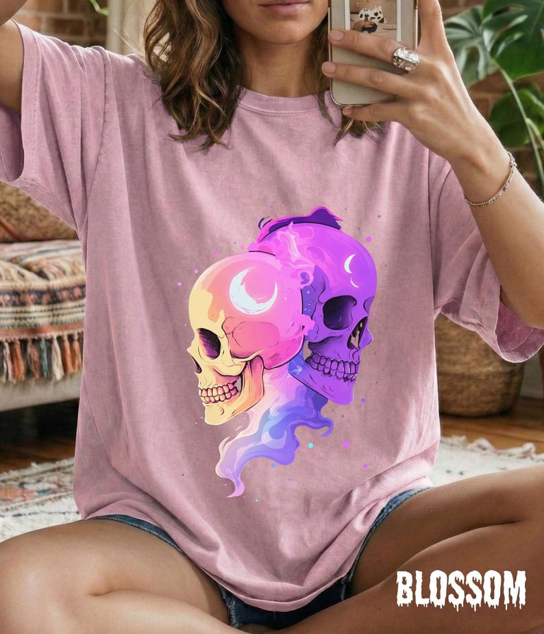 May include: A light pink t-shirt featuring a colorful graphic of two skulls with crescent moon details. The skulls are in shades of yellow, pink, and purple, with a flowing design. The word "BLOSSOM" is printed in a white, stylized font.