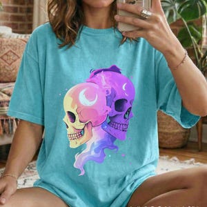May include: A turquoise blue t-shirt featuring a graphic of two skulls with crescent moon details. The skulls are in shades of yellow, pink, and purple. The text "LAGOON BLUE" is at the bottom of the shirt.