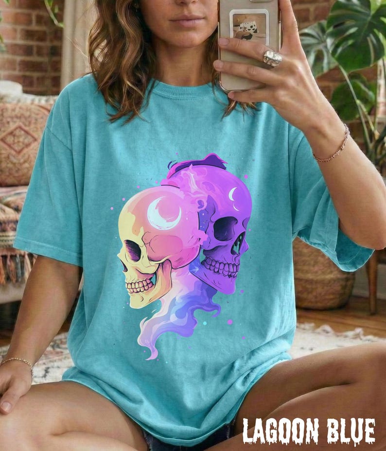 May include: A turquoise blue t-shirt featuring a graphic of two skulls with crescent moon details. The skulls are in shades of yellow, pink, and purple. The text "LAGOON BLUE" is at the bottom of the shirt.