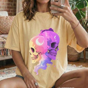 May include: Butter-colored t-shirt featuring a graphic of two stylized skulls, one pink and yellow, the other purple. The skulls have crescent moon details and are surrounded by colorful smoke-like designs. The word "BUTTER" is printed at the bottom.