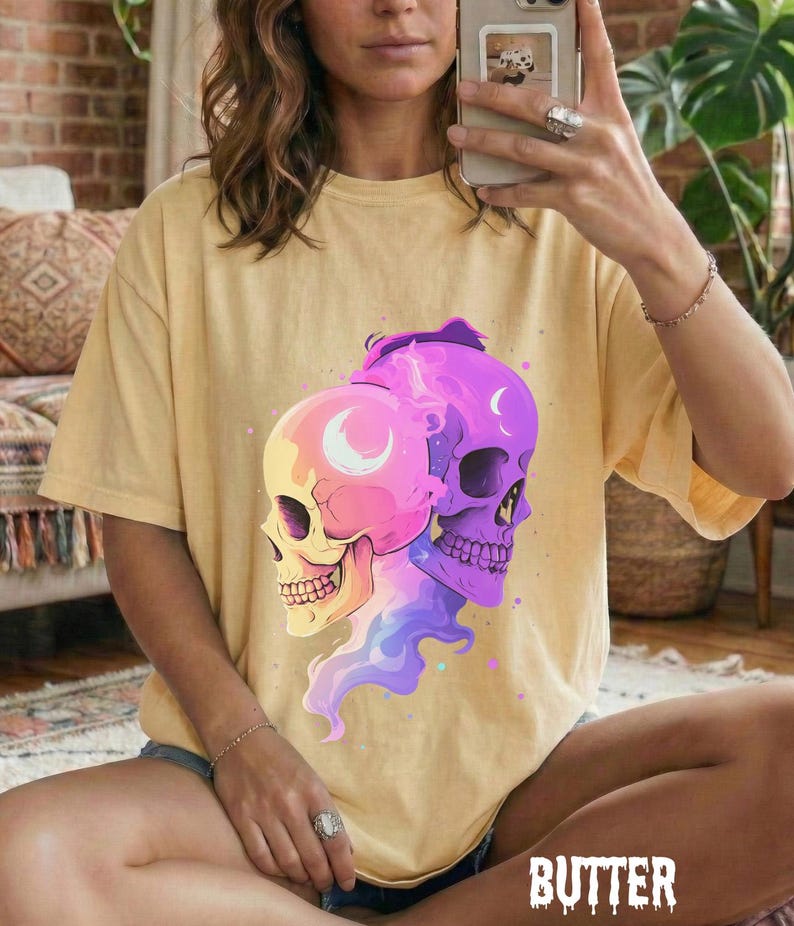 May include: Butter-colored t-shirt featuring a graphic of two stylized skulls, one pink and yellow, the other purple. The skulls have crescent moon details and are surrounded by colorful smoke-like designs. The word "BUTTER" is printed at the bottom.