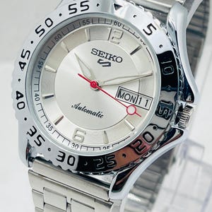 Pode incluir: Um relógio de pulso automático Seiko prateado com mostrador prateado, algarismos pretos e exibição da data. O relógio tem uma pulseira de aço inoxidável e uma moldura giratória com marcações pretas. Os ponteiros são prateados com um ponteiro de segundos vermelho.
