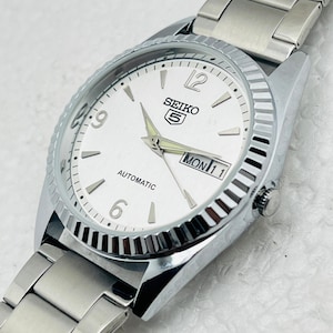 May include: Silver-toned Seiko 5 automatic watch with a white face. The watch features a fluted bezel, silver hands, and a day-date display. The watch band is made of silver metal links.