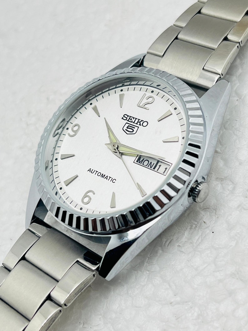 May include: Silver-toned Seiko 5 automatic watch with a white face. The watch features a fluted bezel, silver hands, and a day-date display. The watch band is made of silver metal links.
