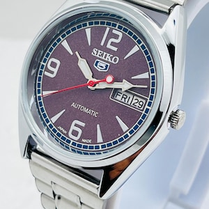 May include: Silver-toned Seiko 5 automatic watch with a dark purple dial. The watch features a stainless steel band, a day-date display, and luminous hands and markers. The watch face displays the number 12, 9, and 6. The watch is made in Japan.
